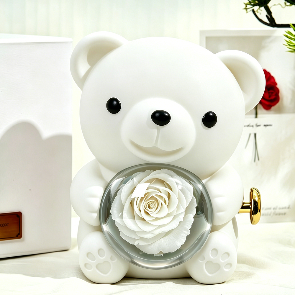 Bloom and Forever Rotating Eternal Rose Bear Gift Box in white, front view showing a white preserved rose encased in a clear heart-shaped acrylic globe held by the bear, with a gold wind-up key on the side, displayed beside a white outer gift box with personalised gold plaque on a cream surface