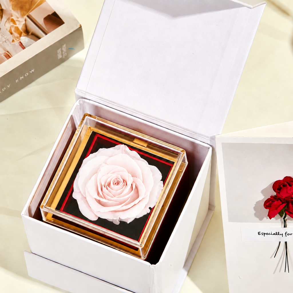 A preserved light pink rose displayed inside a clear square acrylic frame with a gold border and black backing, set in an open white gift box. A matching red flower card is partially visible on the right.