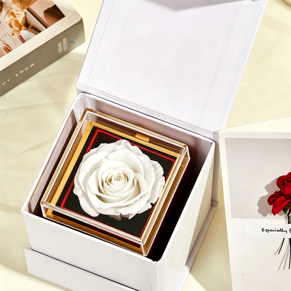 Top-down view of the white Bloom and Forever Rotating Eternal Rose Jewelry Gift Box nestled inside an open white outer gift box, showing a pure white preserved rose under a clear acrylic lid with gold frame on a black base, with an Especially for You greeting card beside it on a cream surface